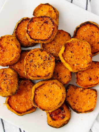 A plate of roasted sweet potato rounds, seasoned with pepper and spices, arranged on a white dish atop a white cloth with black stripes.