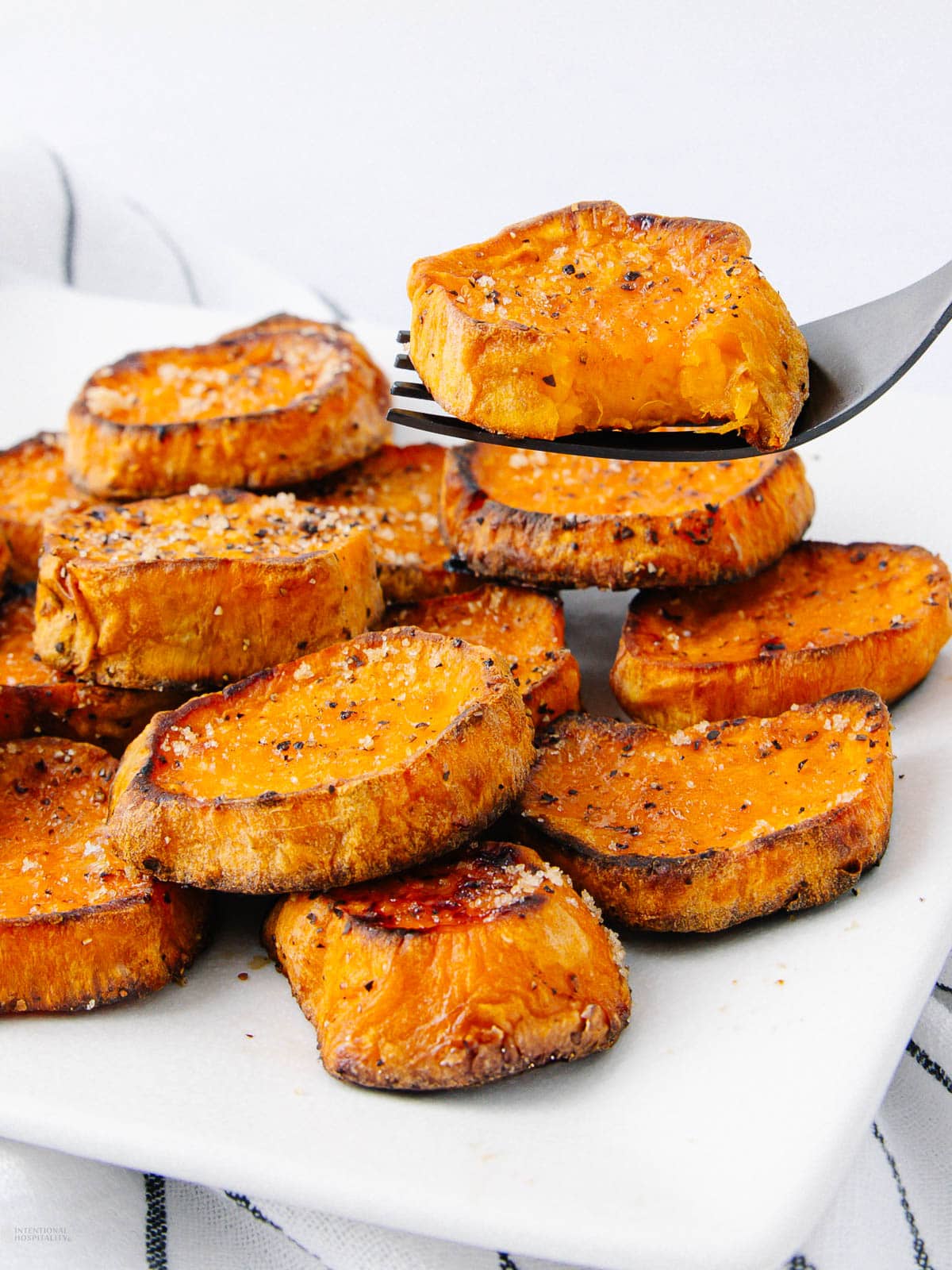A fork holds a slice of roasted sweet potato above a white plate filled with more roasted sweet potato slices, all seasoned with salt and pepper.