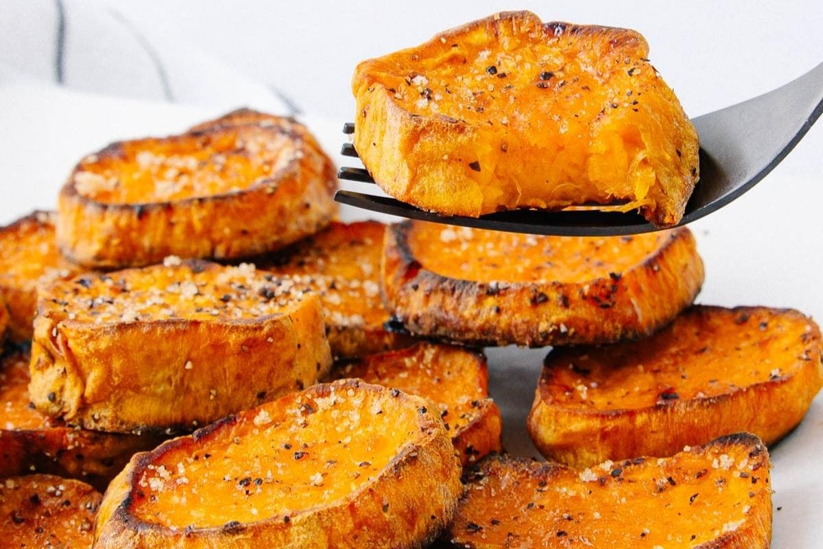 A close-up of roasted sweet potato slices, seasoned with salt and pepper, with one slice held up on a fork above a pile of more slices in the background.