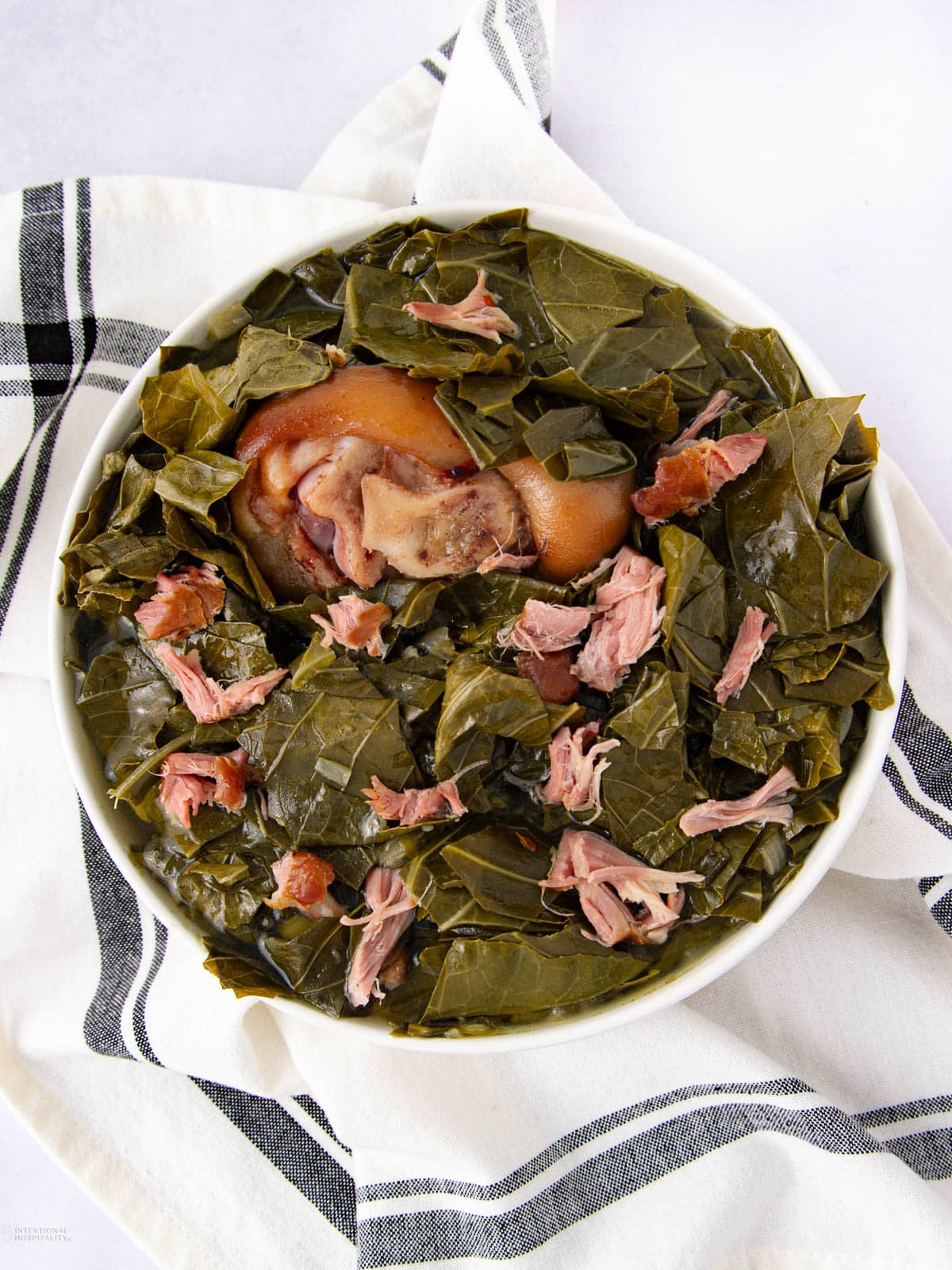 A bowl of cooked collard greens with pieces of smoked ham hock, served on a white towel with black stripes, viewed from above.