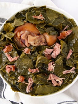A bowl of cooked collard greens with pieces of smoked meat and a large pork hock in the center, set on a white cloth with black stripes.