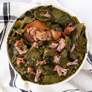 A bowl of cooked collard greens with pieces of smoked meat and a large pork hock in the center, set on a white cloth with black stripes.