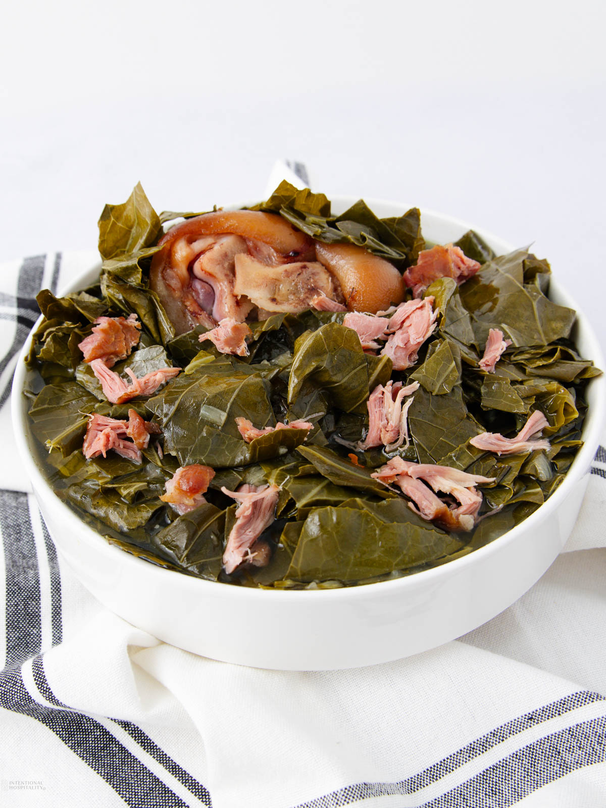 A white bowl filled with cooked collard greens and pieces of smoked ham hock, set on a white and black striped cloth against a white background.