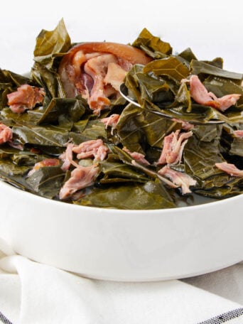 A white bowl filled with cooked collard greens and pieces of pork, set on a white and black striped cloth with a silver spoon resting in the bowl.