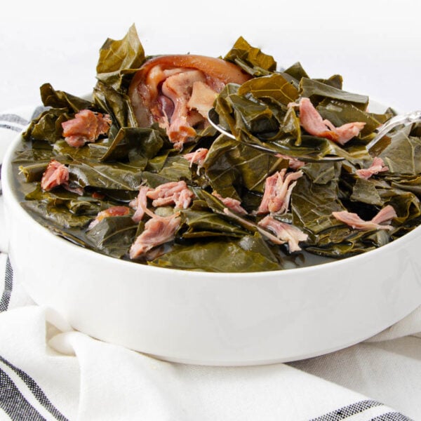 A white bowl filled with cooked collard greens and pieces of pork, set on a white and black striped cloth with a silver spoon resting in the bowl.