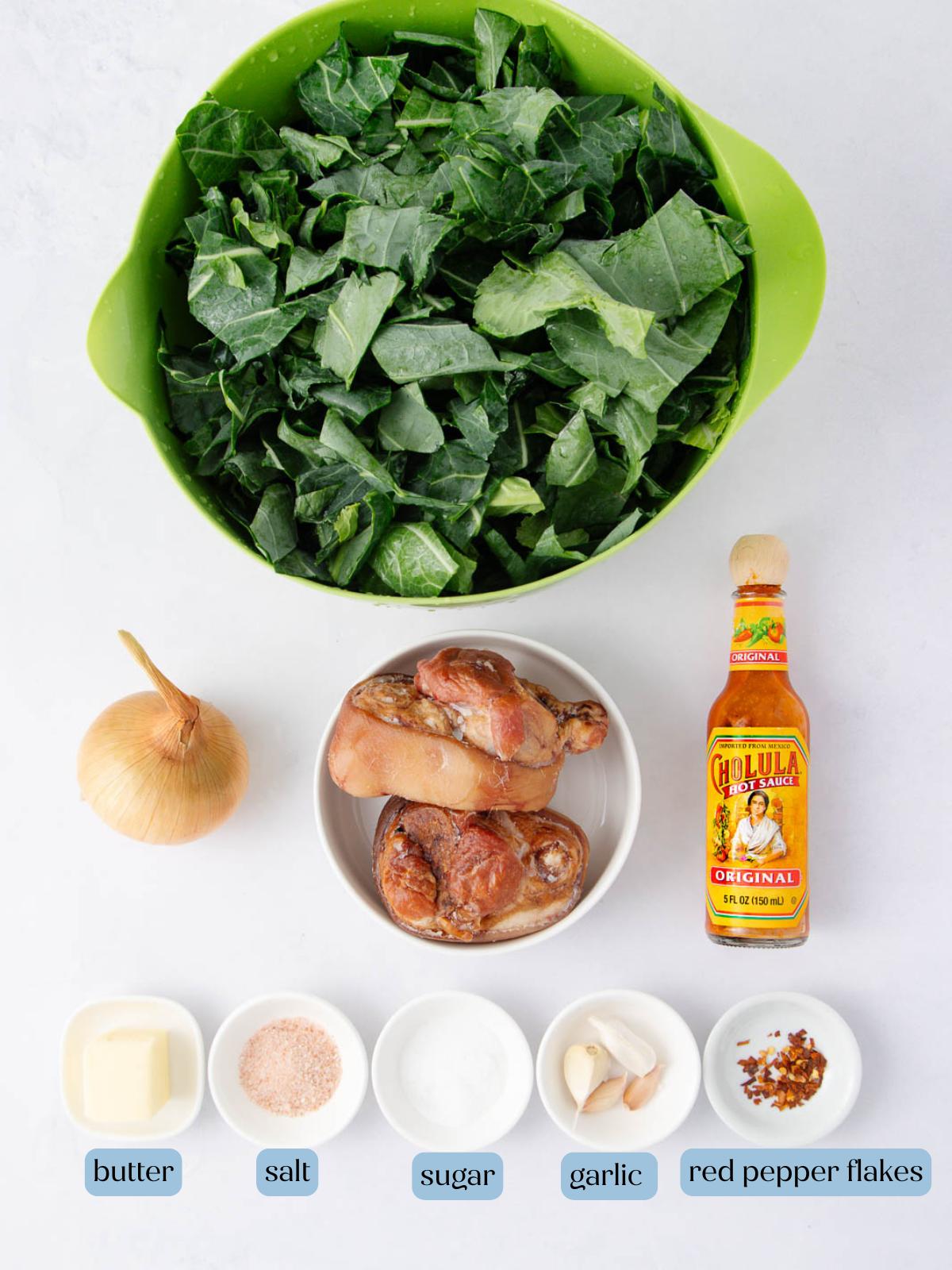 A bowl of chopped collard greens, a whole onion, smoked ham hocks, hot sauce, butter, salt, sugar, garlic cloves, and red pepper flakes are arranged on a white surface.