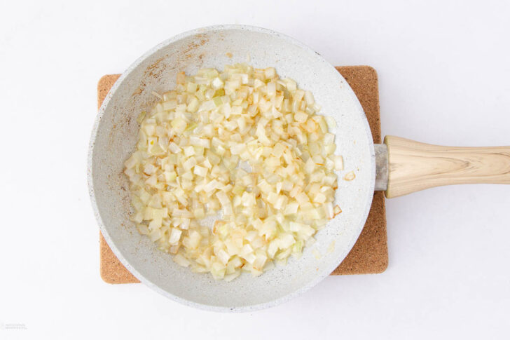 A white frying pan with chopped onions sautéing until golden, placed on a brown cork trivet against a white background.