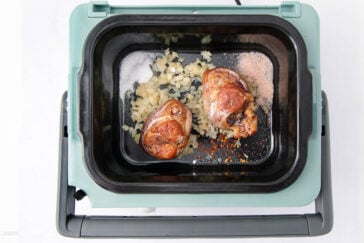 Two browned pork hocks sit on top of chopped onions, salt, and spices in the black insert of a slow cooker with a mint green exterior, ready for cooking.