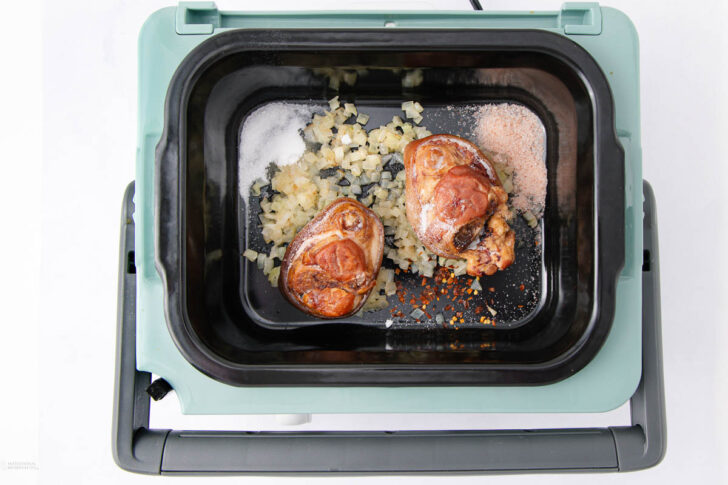 Two browned pork hocks sit on top of chopped onions, salt, and spices in the black insert of a slow cooker with a mint green exterior, ready for cooking.