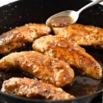 Golden-brown chicken tenders cooking in a skillet, with a spoon drizzling a glossy sauce over the pieces. The chicken is seasoned and sizzling in the sauce.