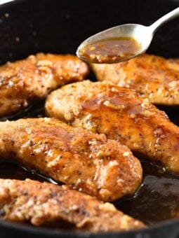 Golden-brown chicken tenders cooking in a skillet, with a spoon drizzling a glossy sauce over the pieces. The chicken is seasoned and sizzling in the sauce.