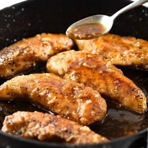 Golden-brown chicken tenders cooking in a skillet, with a spoon drizzling a glossy sauce over the pieces. The chicken is seasoned and sizzling in the sauce.