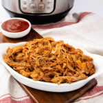 A white dish filled with shredded barbecue pulled pork sits on a wooden board, with a bowl of red barbecue sauce and an Instant Pot in the background. The setting is on a striped towel.