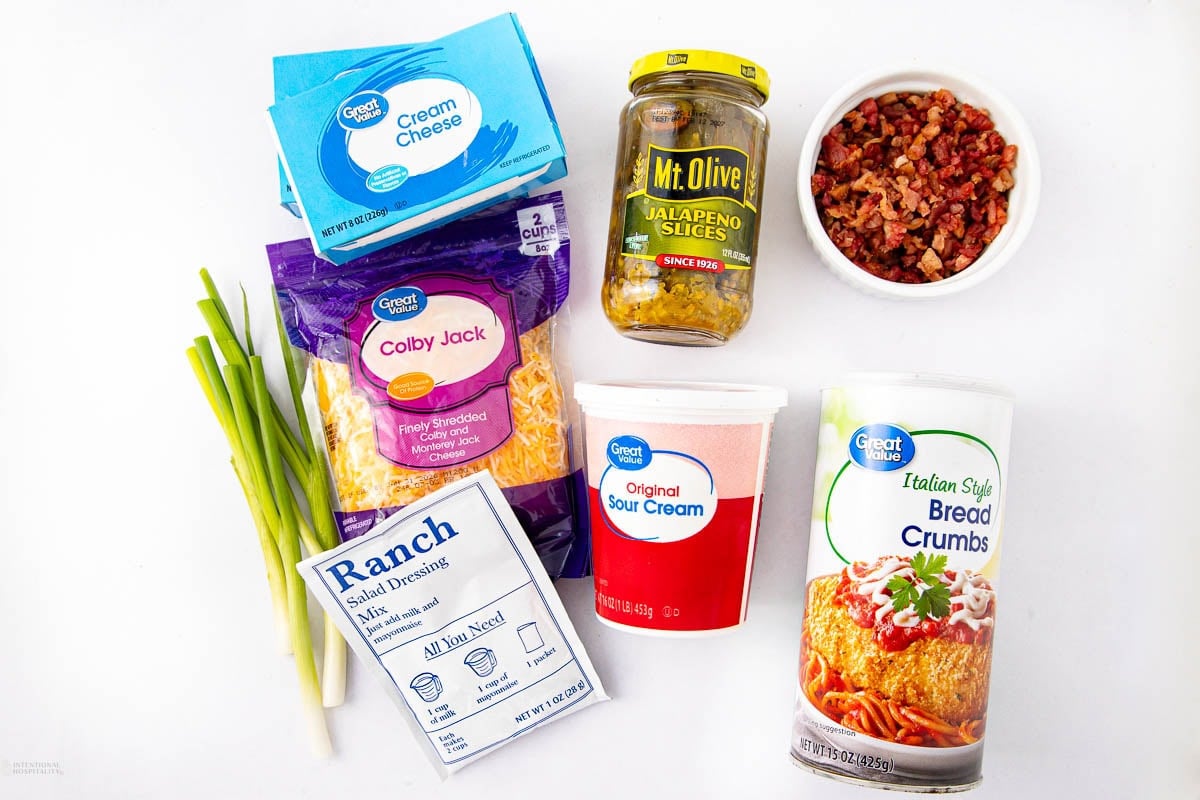 Assorted ingredients on a white surface: cream cheese, shredded Colby Jack cheese, ranch mix, green onions, jalapeño slices, cooked bacon bits, sour cream, and Italian style bread crumbs.