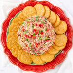 A cheese ball coated in chopped red and green peppers and bacon sits on a decorative red plate, surrounded by a ring of round crackers.