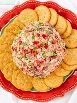 A cheese ball coated in chopped red and green peppers and bacon sits on a decorative red plate, surrounded by a ring of round crackers.