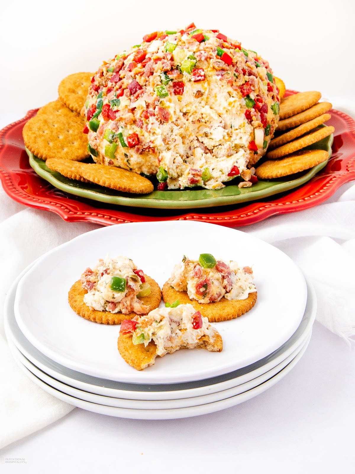 A festive cheese ball coated with colorful diced peppers and bacon sits on a platter, surrounded by round crackers. Three crackers topped with the cheese spread are served on stacked white plates in the foreground.