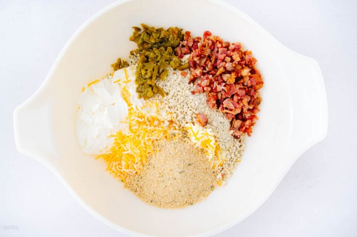 A white mixing bowl containing sour cream, shredded cheese, bread crumbs, chopped bacon, and chopped green chilies, arranged in separate piles on a white background.