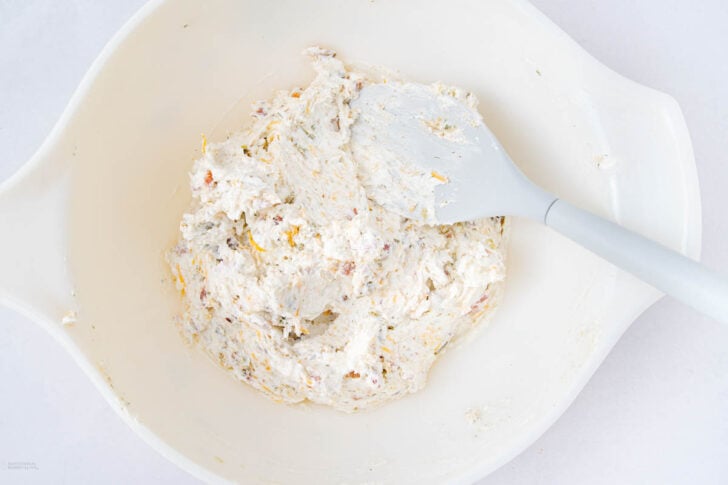 A white mixing bowl with creamy, white cheese mixture and herbs being stirred with a gray spatula.