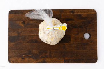 A ball of dough wrapped in a plastic bag and sealed with a yellow clip sits on a dark wooden cutting board with a round hole, against a white background.