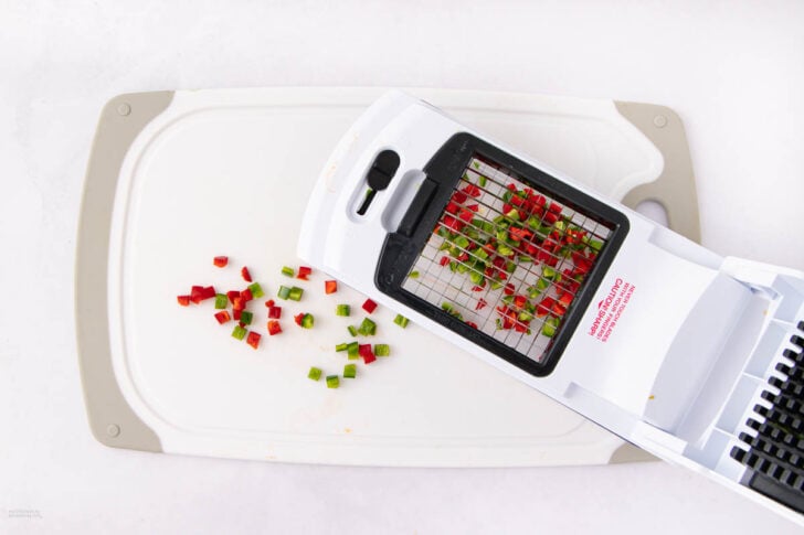 A white vegetable chopper with diced red and green bell peppers sits on a white and gray cutting board, with chopped pepper pieces scattered nearby.