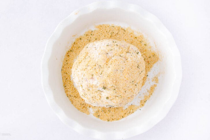 A ball of food, coated in breadcrumbs, sits in a white dish filled with more breadcrumbs on a white background.