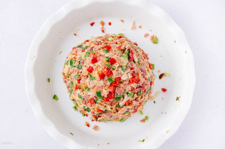 A round cheese ball coated with chopped red and green bell peppers and herbs, placed in a white dish on a white background.