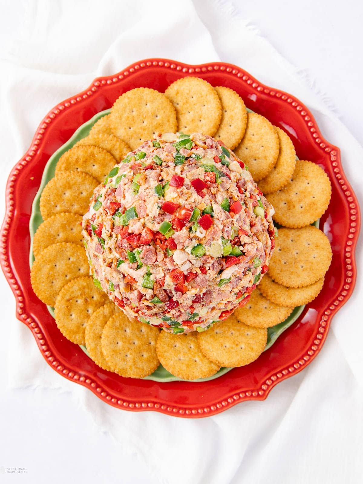 A round cheese ball coated with chopped peppers and nuts is surrounded by a ring of round crackers on a red scalloped plate atop a white cloth.