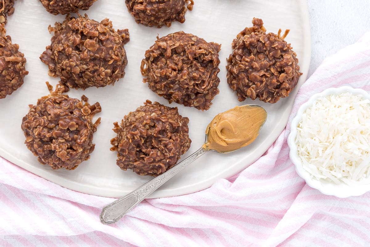 A plate of no-bake chocolate oatmeal cookies sits next to a spoonful of peanut butter and a small bowl of shredded coconut, on a pink-striped cloth.