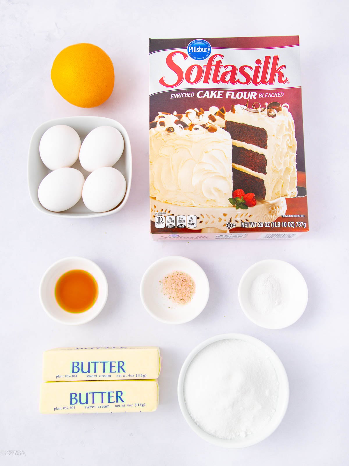 A box of Pillsbury Softasilk cake flour, four eggs in a bowl, an orange, two sticks of butter, a bowl of sugar, vanilla extract, baking powder, and a small bowl of salt arranged on a white surface.