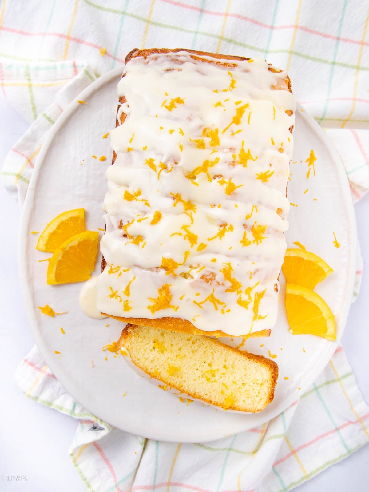 A rectangular loaf of orange cake topped with white icing and orange zest sits on a white plate, garnished with orange slices. One slice is cut and slightly separated from the loaf. A pastel plaid cloth is underneath.