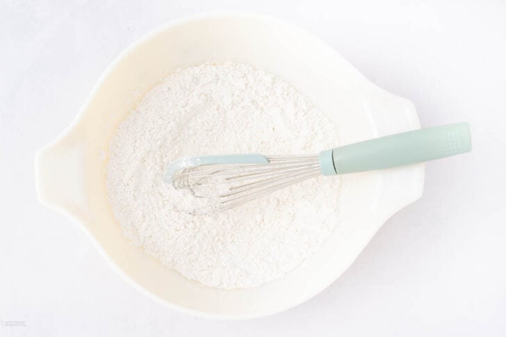 A white mixing bowl filled with flour and a light blue whisk resting on top, viewed from above on a white surface.