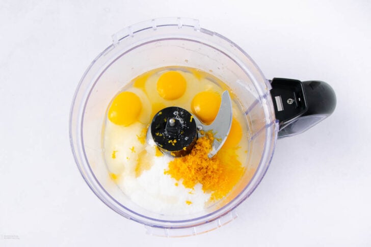 A food processor bowl containing four eggs, sugar, and fresh orange zest on a white surface, viewed from above.