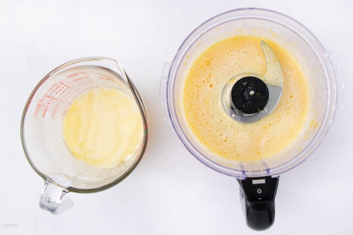 Top-down view of a blender filled with a frothy yellow liquid next to a glass measuring cup containing a pale yellow mixture, both on a white surface.