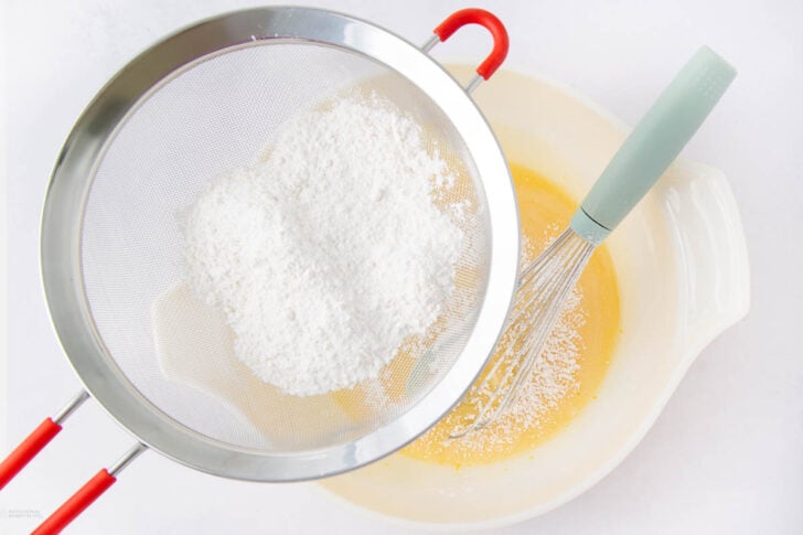 A metal sieve holding powdered sugar is positioned over a bowl with a whisk and a yellow batter, as the sugar is being sifted into the mixture on a white background.