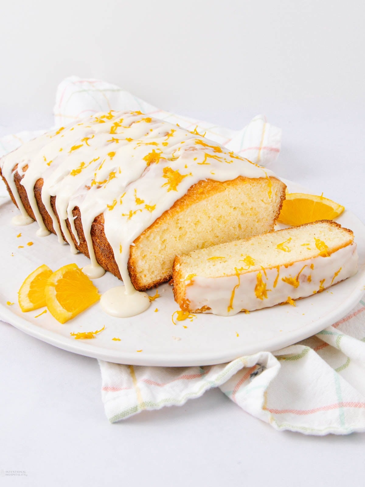 A loaf of orange cake with white icing, garnished with orange zest, sits on a white plate. One slice is cut and laid in front, with orange slices and zest scattered nearby. A white cloth is in the background.