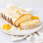 A rectangular loaf of orange cake with white icing and orange zest on top sits on a white plate, with slices of orange and a piece of cake cut from the loaf. A light plaid cloth is underneath the plate.