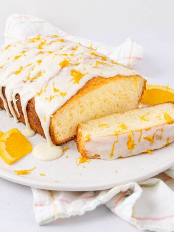 A rectangular loaf of orange cake with white icing and orange zest on top sits on a white plate, with slices of orange and a piece of cake cut from the loaf. A light plaid cloth is underneath the plate.