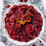 A white bowl filled with chunky cranberry sauce, garnished with grated orange zest, sits on a patterned black and white cloth. A spoon rests in the bowl, partially submerged in the sauce.