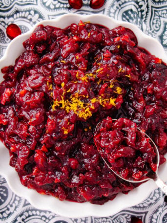 A white bowl filled with chunky cranberry sauce, garnished with grated orange zest, sits on a patterned black and white cloth. A spoon rests in the bowl, partially submerged in the sauce.
