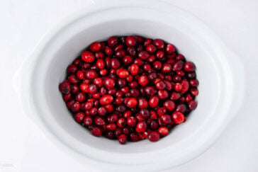 A white oval slow cooker filled with fresh, whole cranberries on a white background.