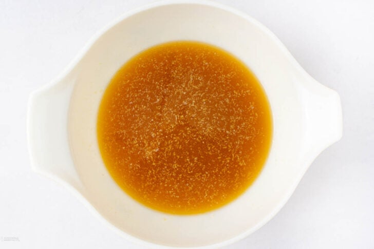 A white bowl filled with a light brown, translucent liquid broth or stock, shown from above on a plain white background.