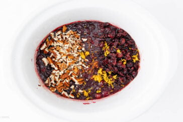 A white slow cooker filled with cranberry sauce, topped with chopped pecans, dried cranberries, and orange zest, arranged in three sections on the surface.