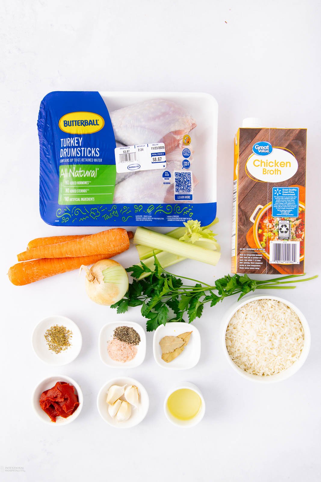 Top-down view of ingredients for a recipe, including packaged turkey drumsticks, chicken broth, carrots, onion, parsley, rice, garlic, tomato paste, oil, and seasonings in small bowls on a white surface.