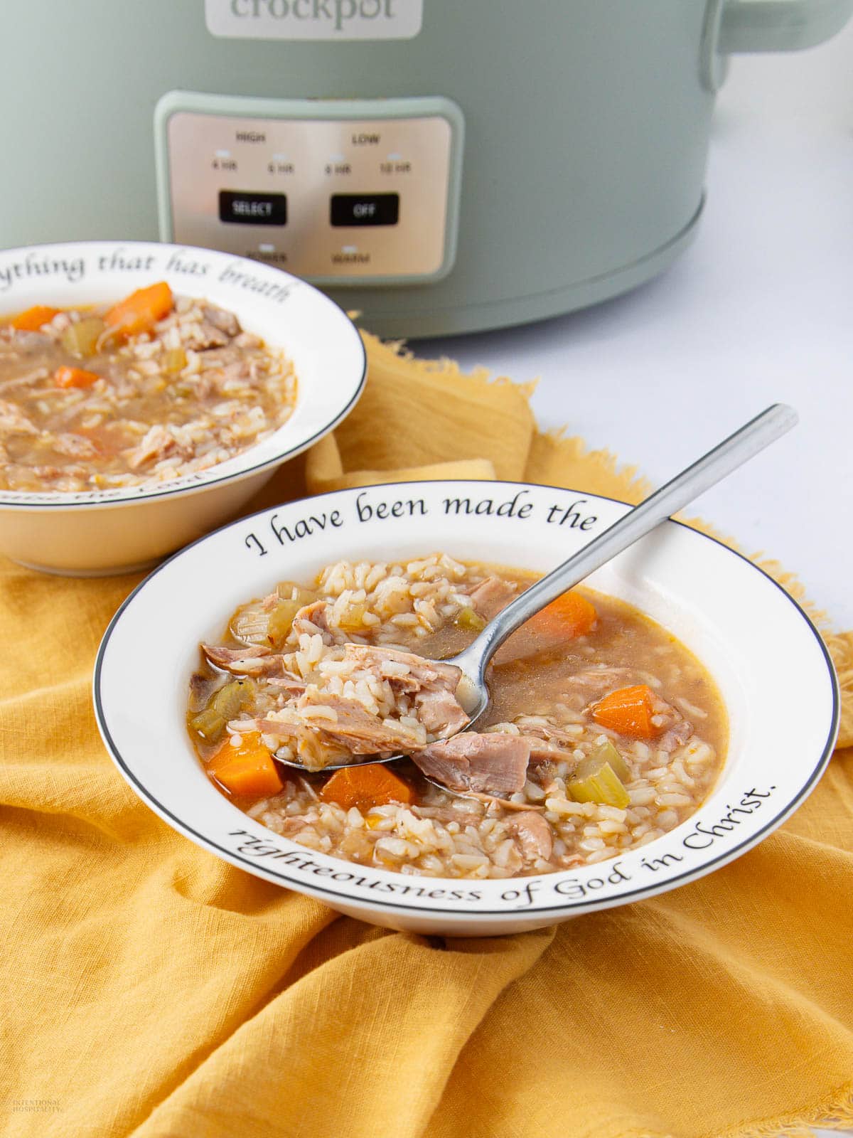 A bowl of soup with rice, vegetables, and shredded meat sits on a yellow cloth, with a spoon resting inside. The bowl has text around the rim and a slow cooker is in the background.