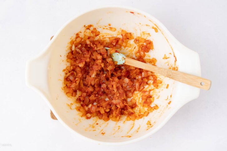 A white mixing bowl filled with sautéed diced onions and tomato paste, with a spatula resting inside, sitting on a white surface.