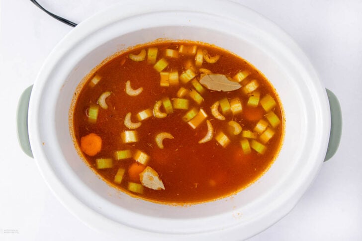A slow cooker filled with a tomato-based broth, diced celery, sliced carrots, and two bay leaves, viewed from above on a white background.