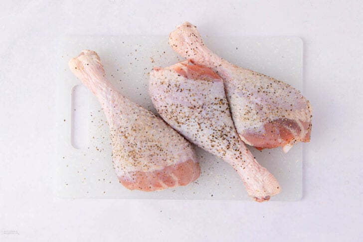 Three raw turkey legs seasoned with salt and black pepper, placed on a white cutting board with a handle, on a light-colored surface.