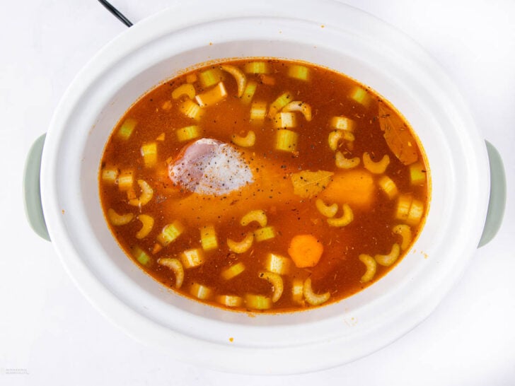 A white slow cooker filled with broth, chopped celery, carrot slices, a bay leaf, and a piece of seasoned raw meat, viewed from above on a white background.