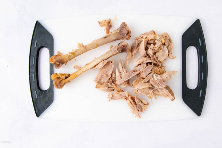 A white cutting board with black handles holds two bare turkey bones and a pile of shredded cooked turkey meat on a white surface.